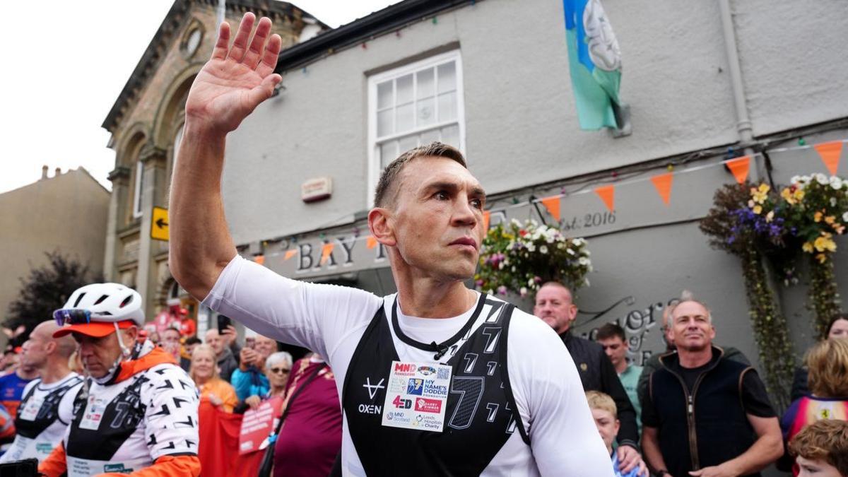 A man in a white sports shirt with a black running bib over the top is waving to a crowd of people outside a grey pub that has orange bunting and a Yorkshire flag flying.
