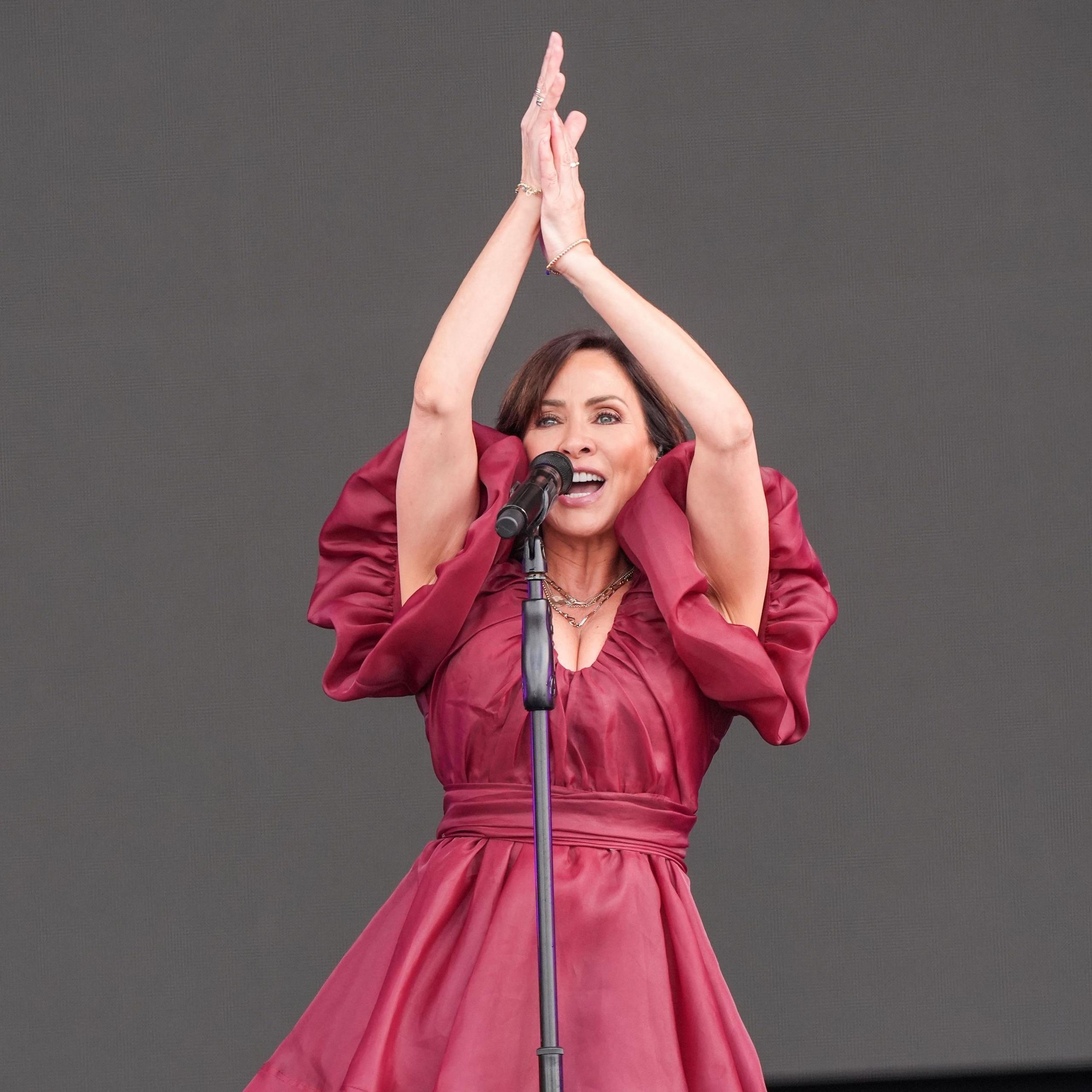 Natalie Imbruglia performs on stage in a cherry red ballgown, with her hands raised above her head as she encourages the audience to clap