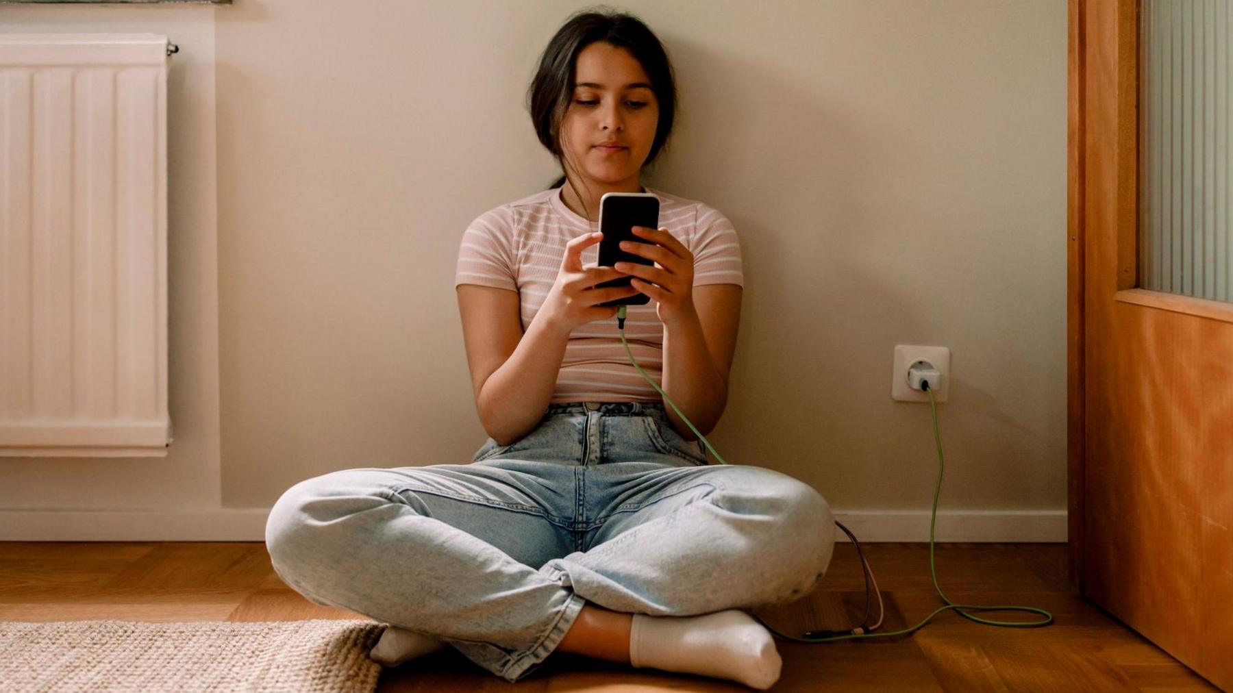 A young girl is seen sat crossed legged on the floor scrolling on a smartphone while it is charging