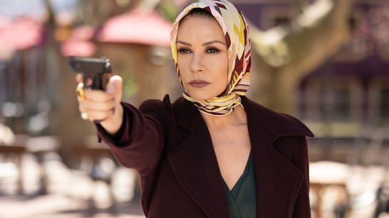Catherine Zeta-Jones holding a gun up, wearing a scarf around her head and a maroon jacket in the Kill Jackie series.