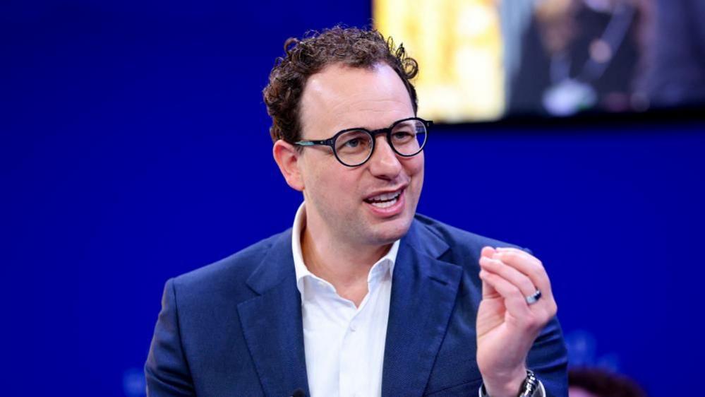 Dario Amodei hld the closed fingers of his left hand together as he mades a point. He is wearing a white shirt and blue jacket, black-rimmed glasses and has slightly curly hair. He is sat against a blue background, with a yellow-lit square just behind and to the left of his head.