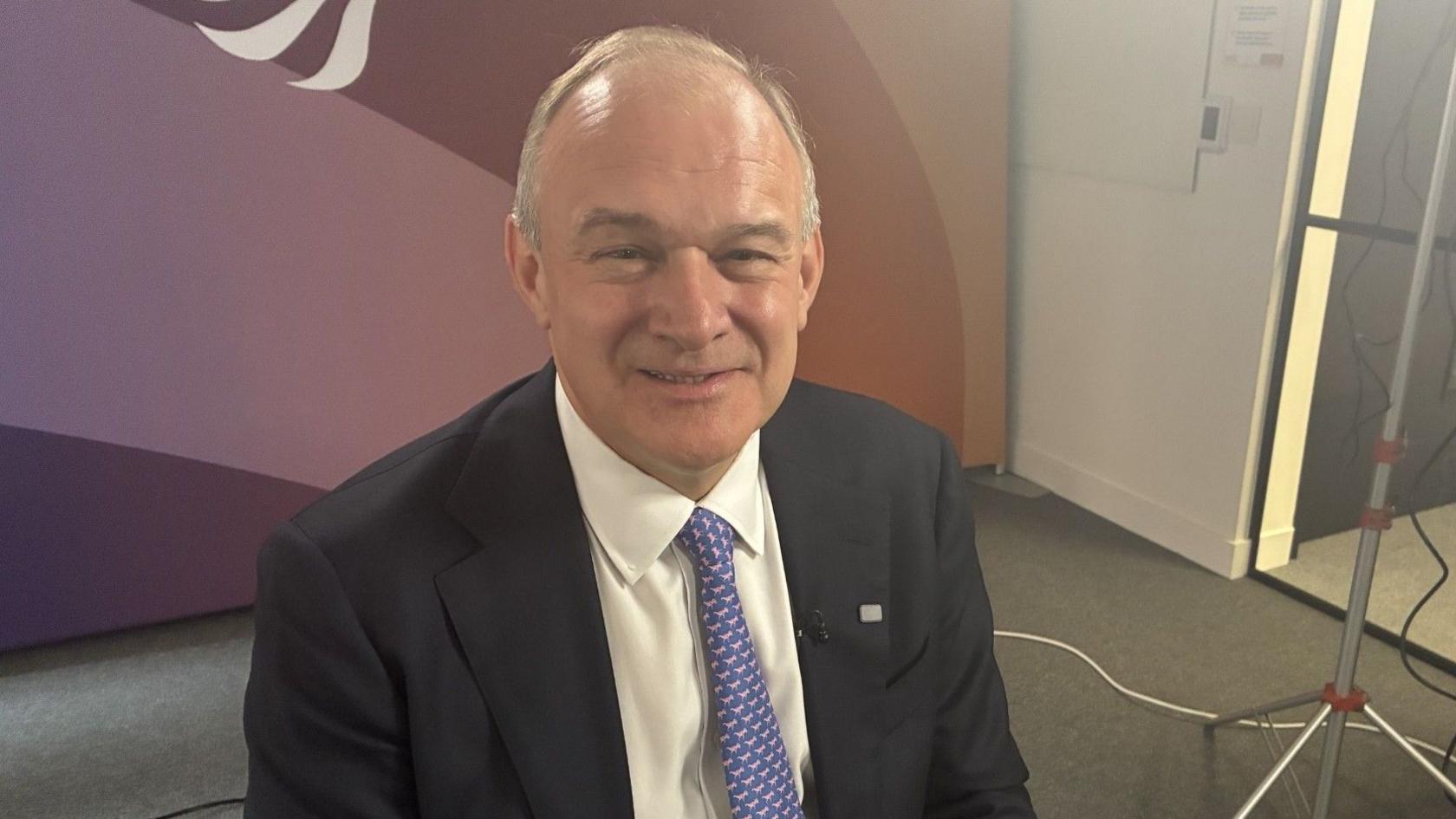 Sir Ed Davey is sitting in a small TV studio. He is smiling, dressed in a suit, white shirt and purple tie, and has short thinning brown and white hair.