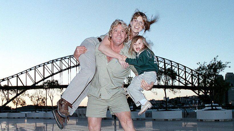 A man wearing khaki shirt and shorts holding a young child with one arm and an adult woman with his other arm with the Sydney Harbour Bridge in the background