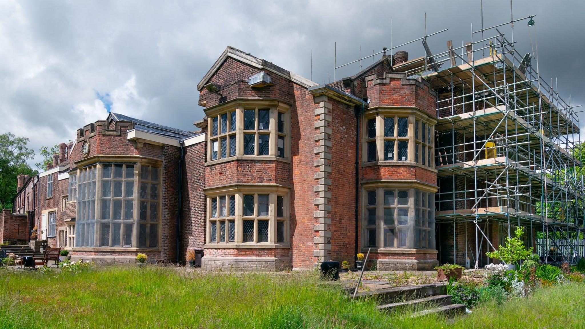 A large manor house has scaffolding on the right hand side. Steps lead up from the lower level of the grounds to the house itself.