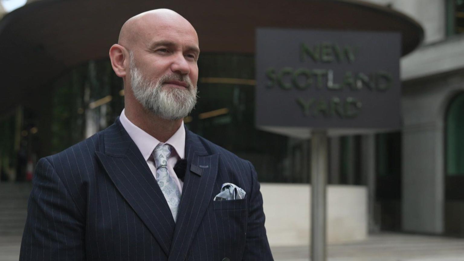Andy Featherstone is standing outside New Scotland Yard, with its spinning sign in the background. He wears a dark coloured pinstripe suit.