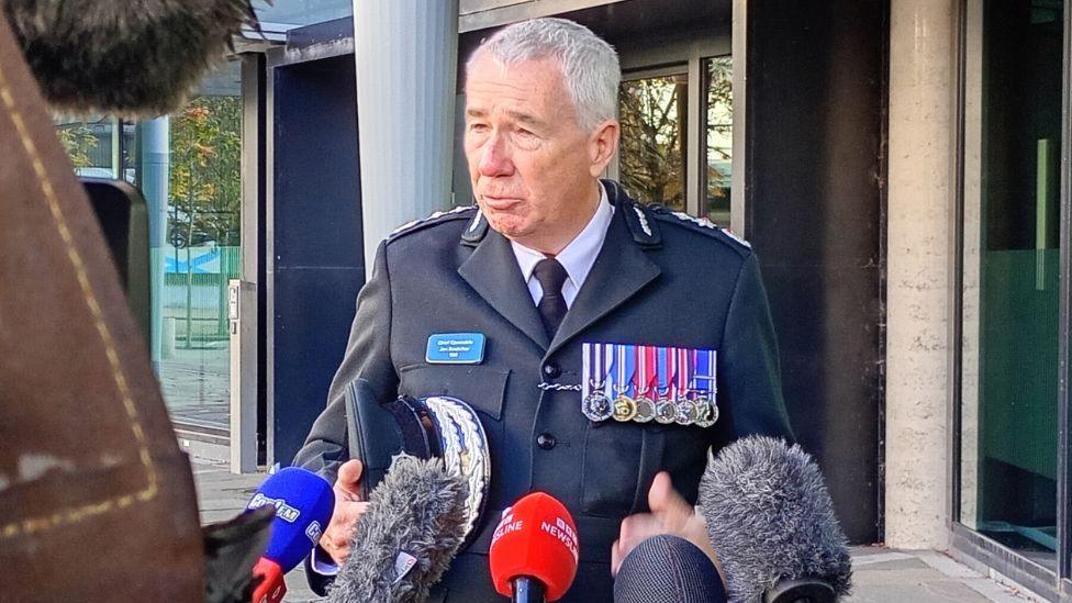 A man in a navy jacket, adorned with medals, stood with numerous microphones in front of him