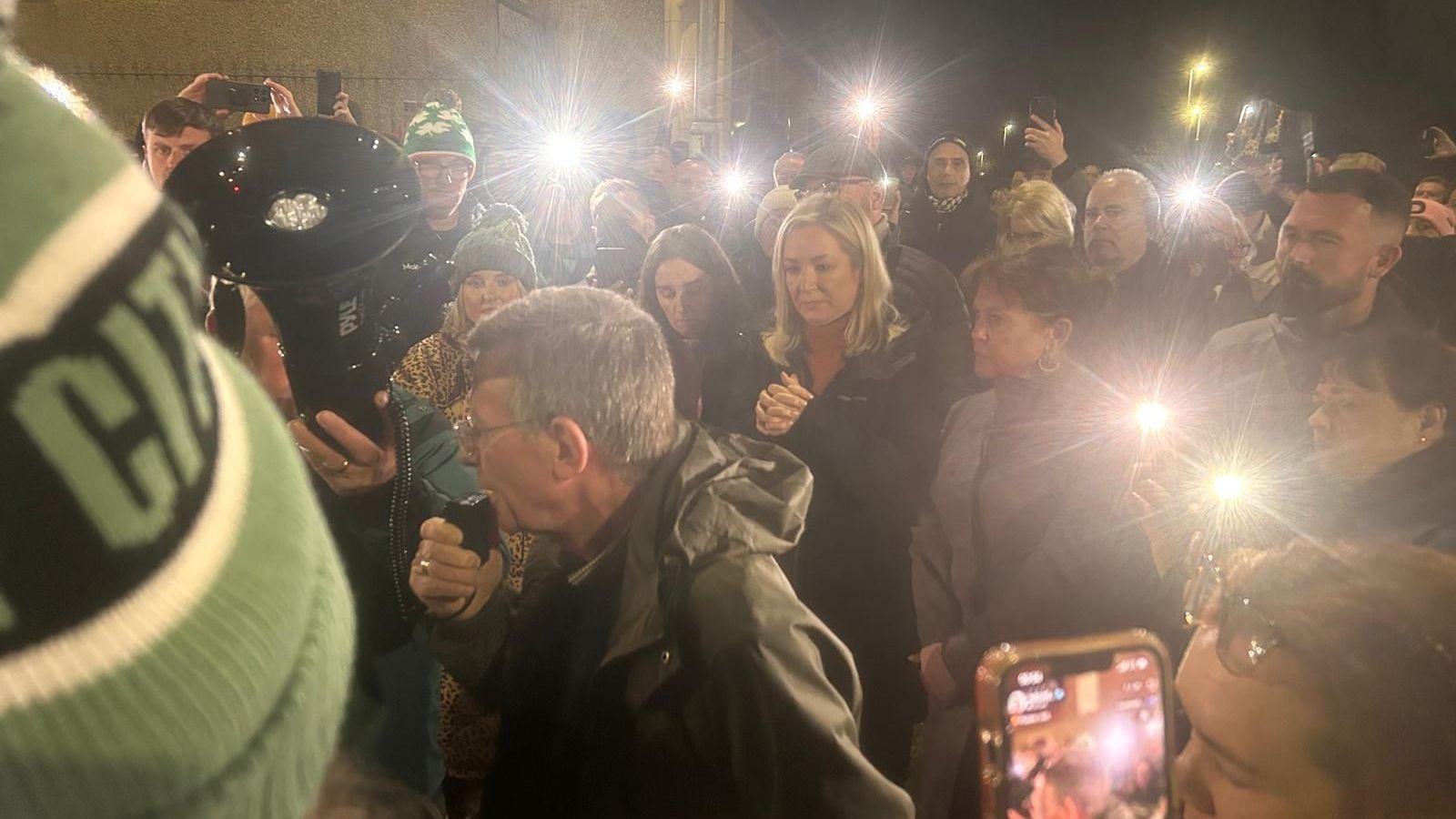 A man with grey hair in an anorak, speaking into a megaphone, surrounded by protesters and politicians