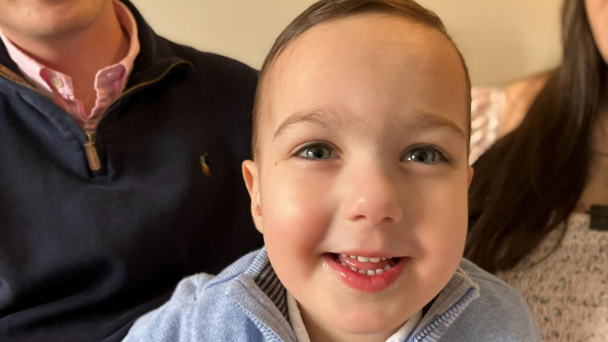 A bright-eyed little boy smiles at the camera. he wears a pale blue jumper. Behind him are sat his parents.