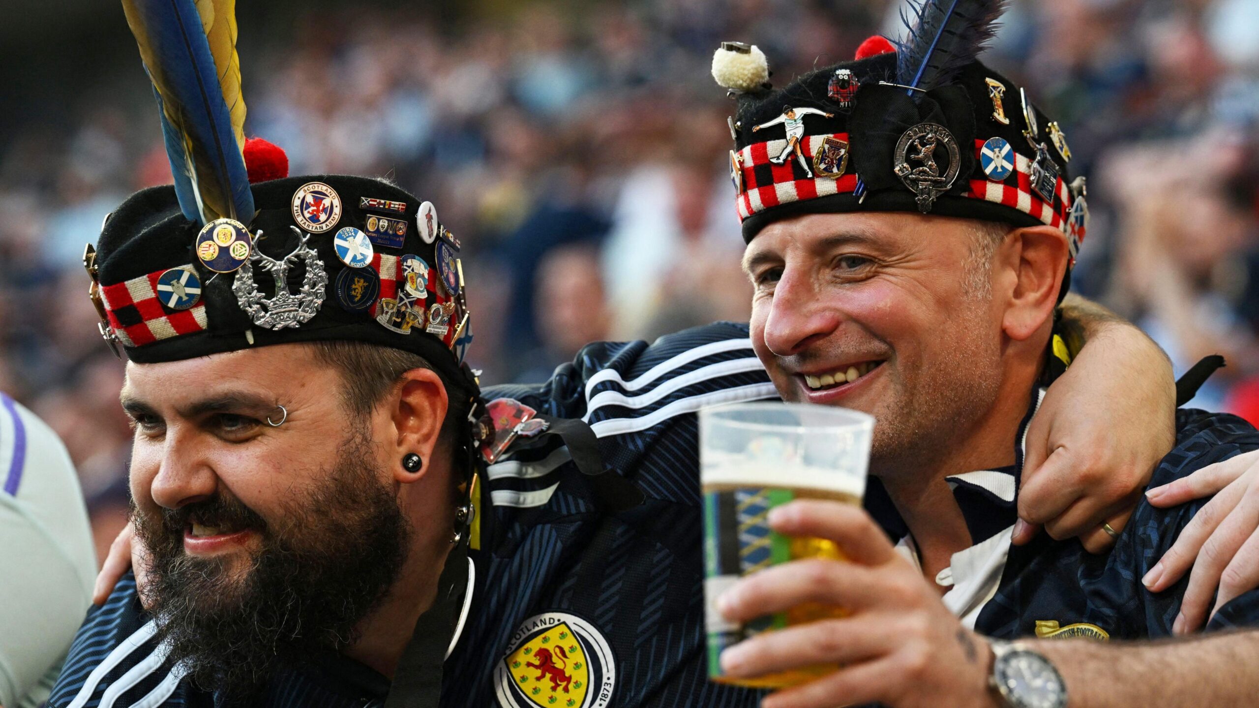 Two Scotland football fans with arms around each other and wearing replica dark blue Scotland tops and bonnets with various lapel pins on it. One of the men has a pint glass with beer in it in one hand.