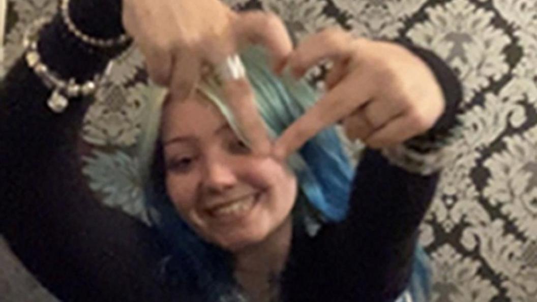 A young girl with long blue hair holds up a heart sign with her hands. She is smiling and wearing a long sleeved black top, silver bracelets. She is sitting in front of black and silver patterned wallpaper.