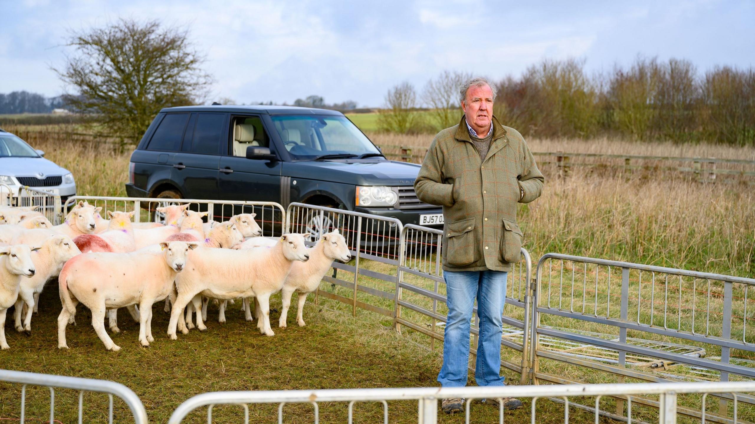 Jeremy Clarkson stood in front of sheep in a holding pen with his hands in his pockets on his farm, a land rover is parked behind him.