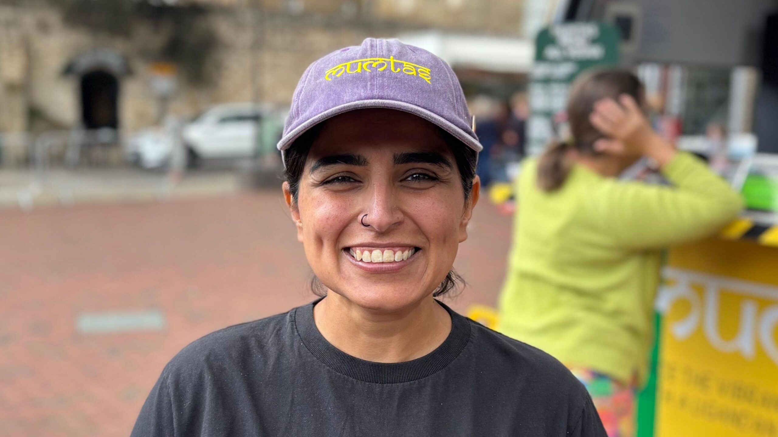 A woman - Tasnime Rotherham - smiling straight at the camera. She is standing in a market square in front of a food truck. She is wearing a black T-shirt and a purple hat with the word 'Mumtas' embroidered in yellow.
