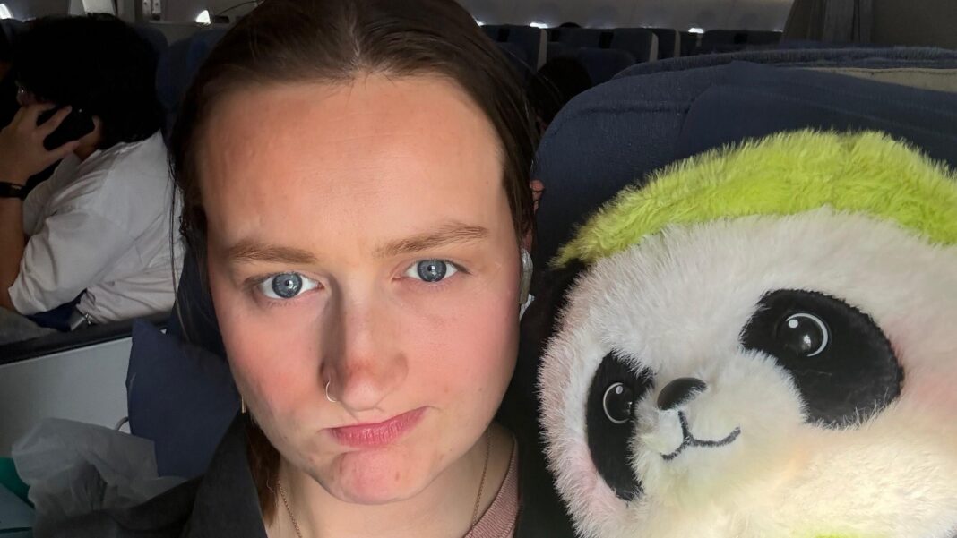 Addie Kitson sits in her airplane seat with a fluffy panda bear which she brought back from China after seeing many of them in China. She is a young woman with brown hair and blue eyes and a nose ring.
