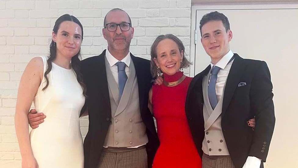 Kate and her husband stand in the middle with their daughter on the far left and son on the far right. They are all dressed smartly and smiling for the camera.
