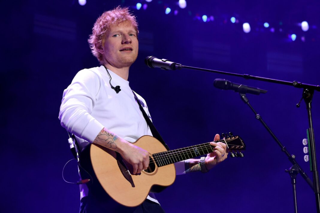 Ed Sheeran looks off to the camera on stage, he is wearing a white jumper and has ginger hair. He is holding a guitar.