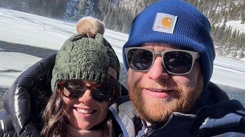 Timby and Mangan both wear winter knit caps, sunglasses and heavy coats in the Colorado snow, a forest treeline visible behind them