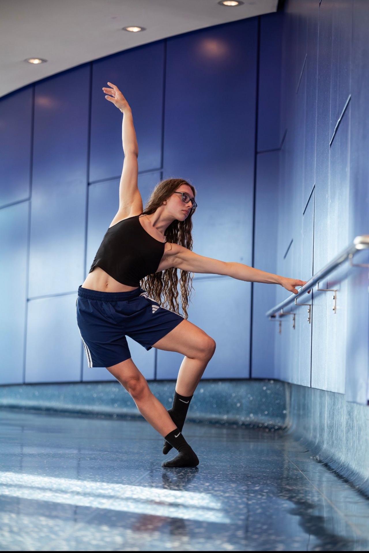 Anna-May seen holding a barre as she dances in a studio while wearing glasses, a black vest top, adidas shorts and socks