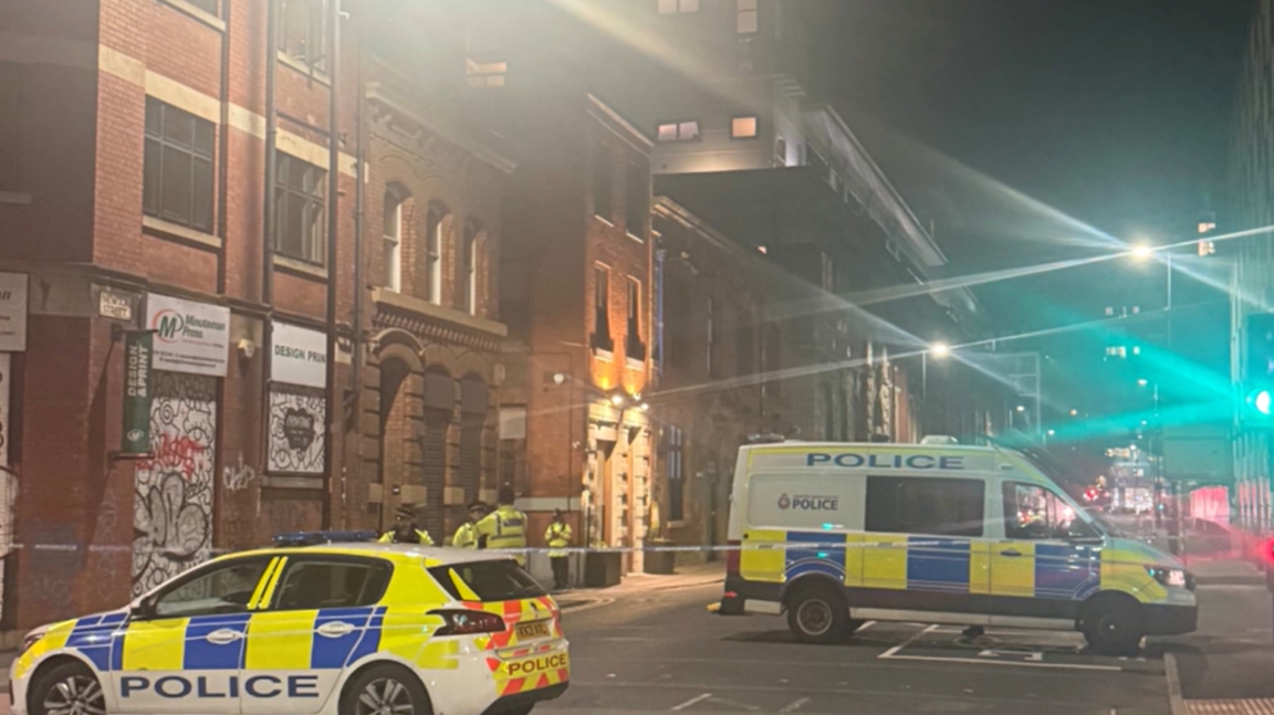 A police car and a van and several officers stand guard by a police cordon in a city-centre location.