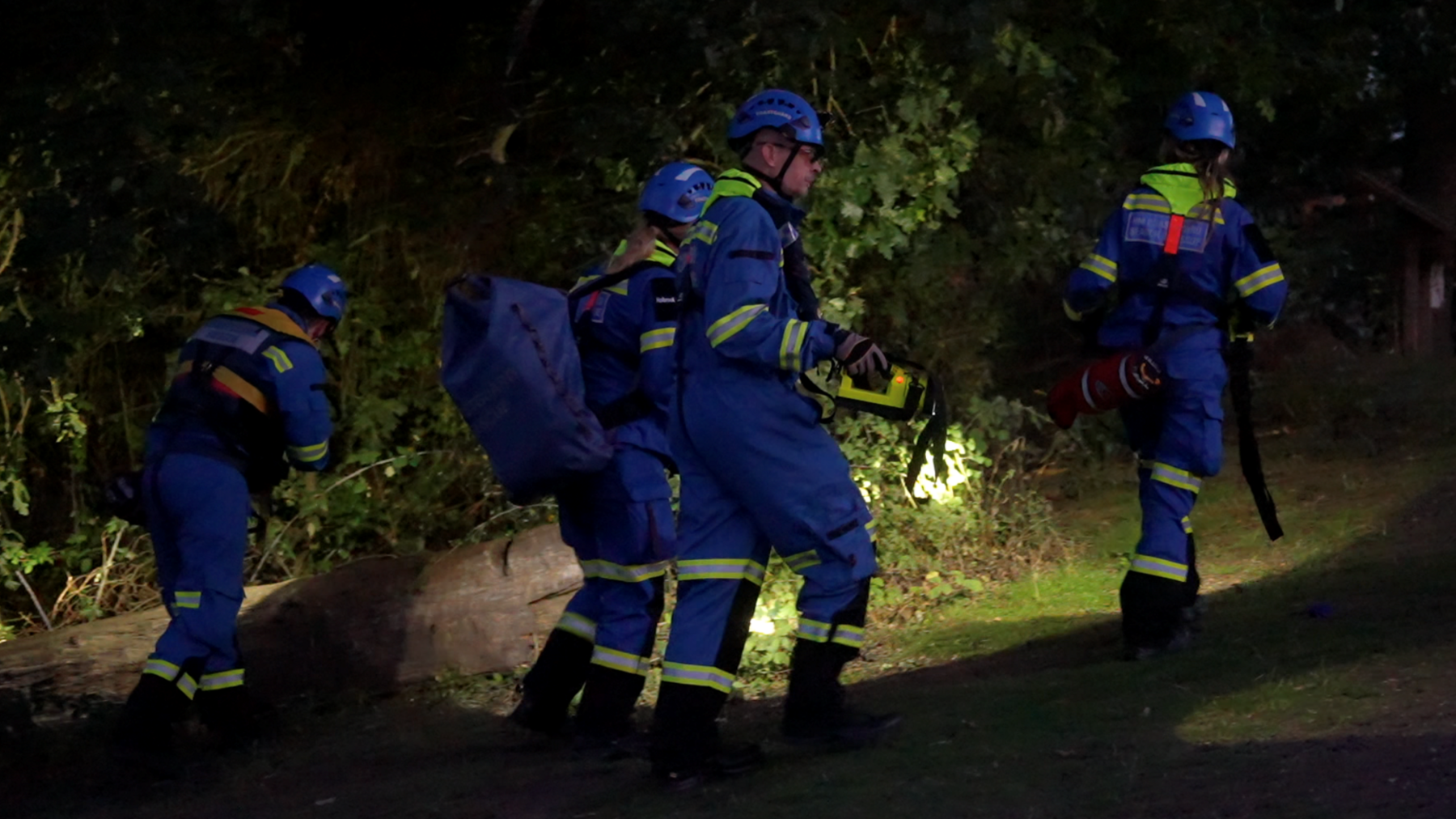 Four members of an emergency search and rescue team use torches to search a wooded area. They are wearing blue helmets and overalls.