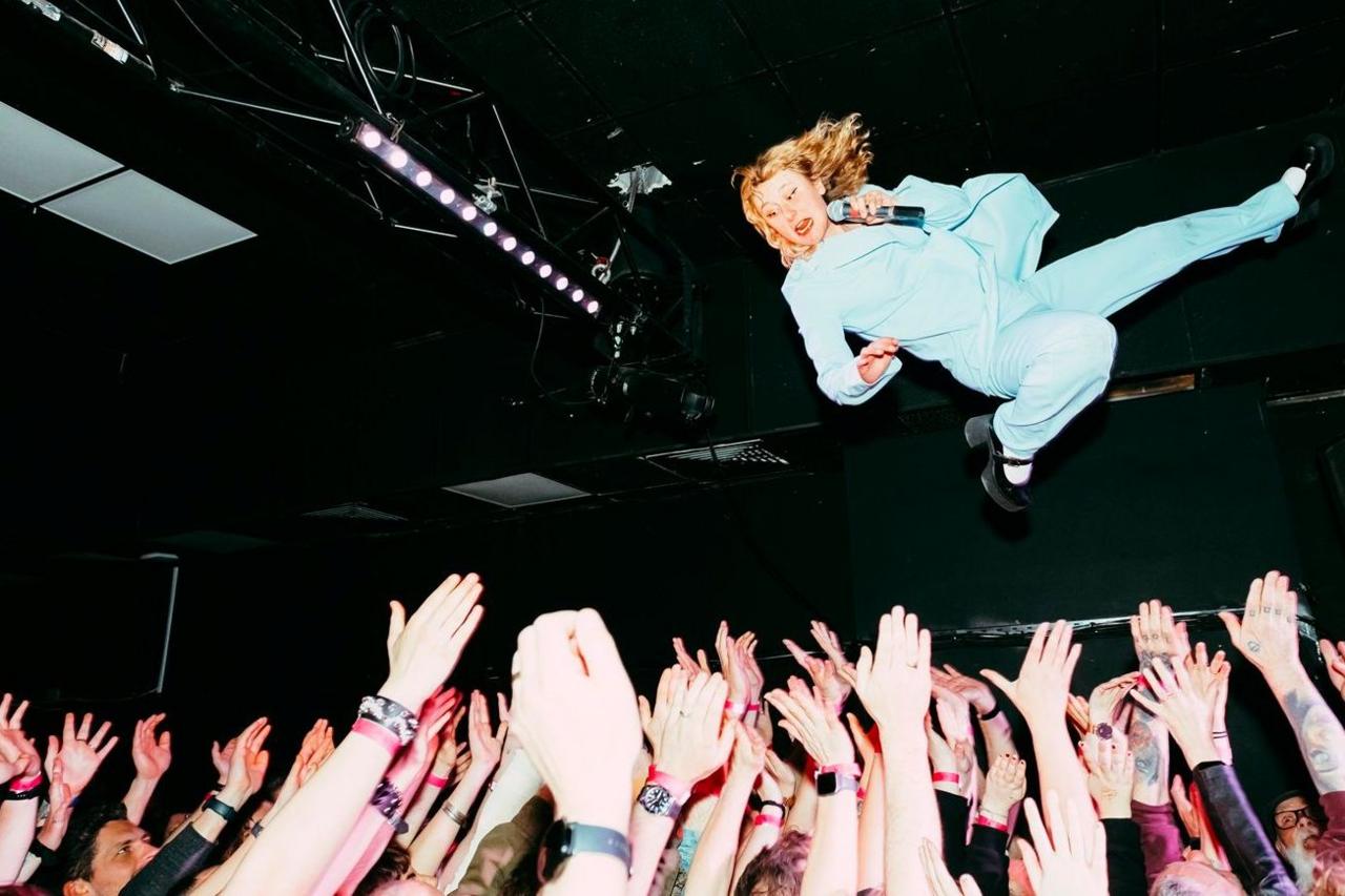 A suited Phoebe Lunny of Lambrini Girls jumping backwards into a sea of hands held aloft in the crowd at a gig