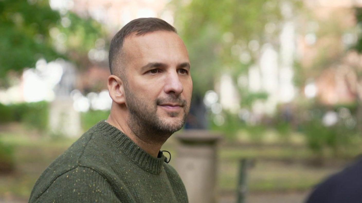 A man with dark hair and heavy stubble, wearing a dark green jumper, seated and being interviewed in a park.
