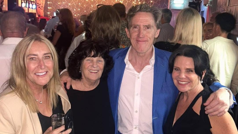 Rob Brydon is wearing a blue blazer and white shirt. He is standing next to the staff members of the club and the stewardess Sarah Prescott. In the background, there is a crowd of people facing the other way.