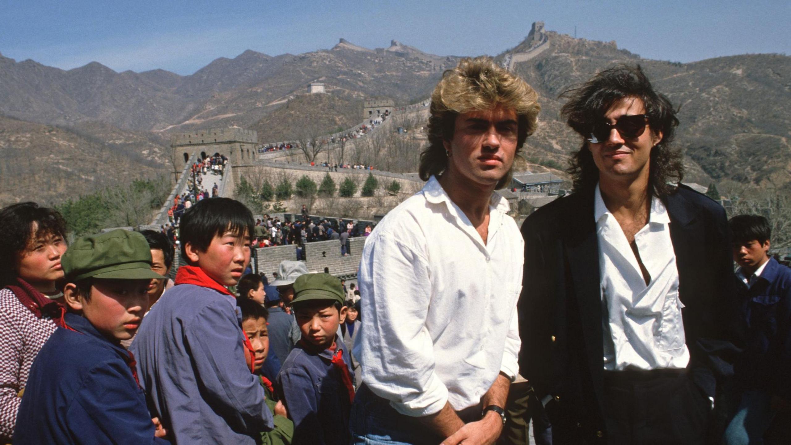 Wham! stand among intrigued children on the Great Wall of China during the band's 1985 visit to the country. George Michael and Andrew Ridgeley have shoulder-length hair and are wearing white shirts. Ridgeley is also wearing a black jacket.