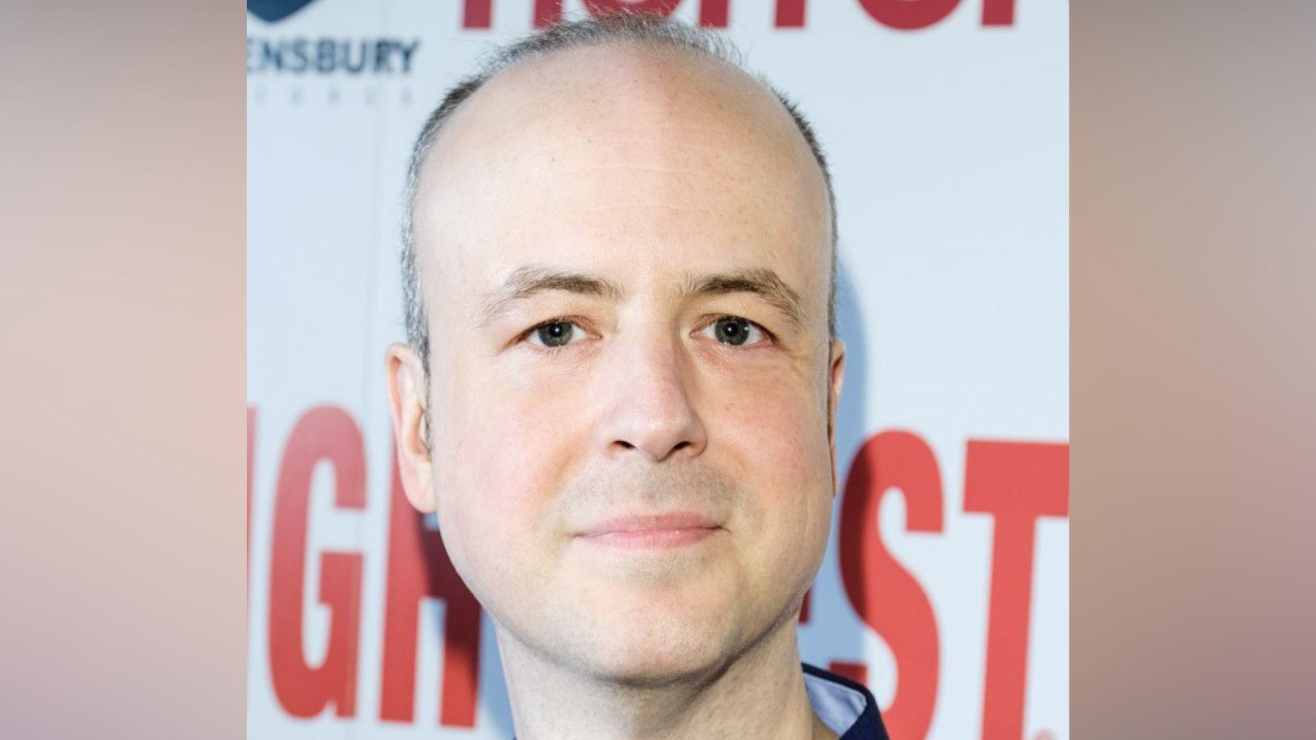 Marcus Hearn has short grey hair and is wearing a navy jumper. He has a light smile and is standing in front of a white wall which has several pieces of branding on it.