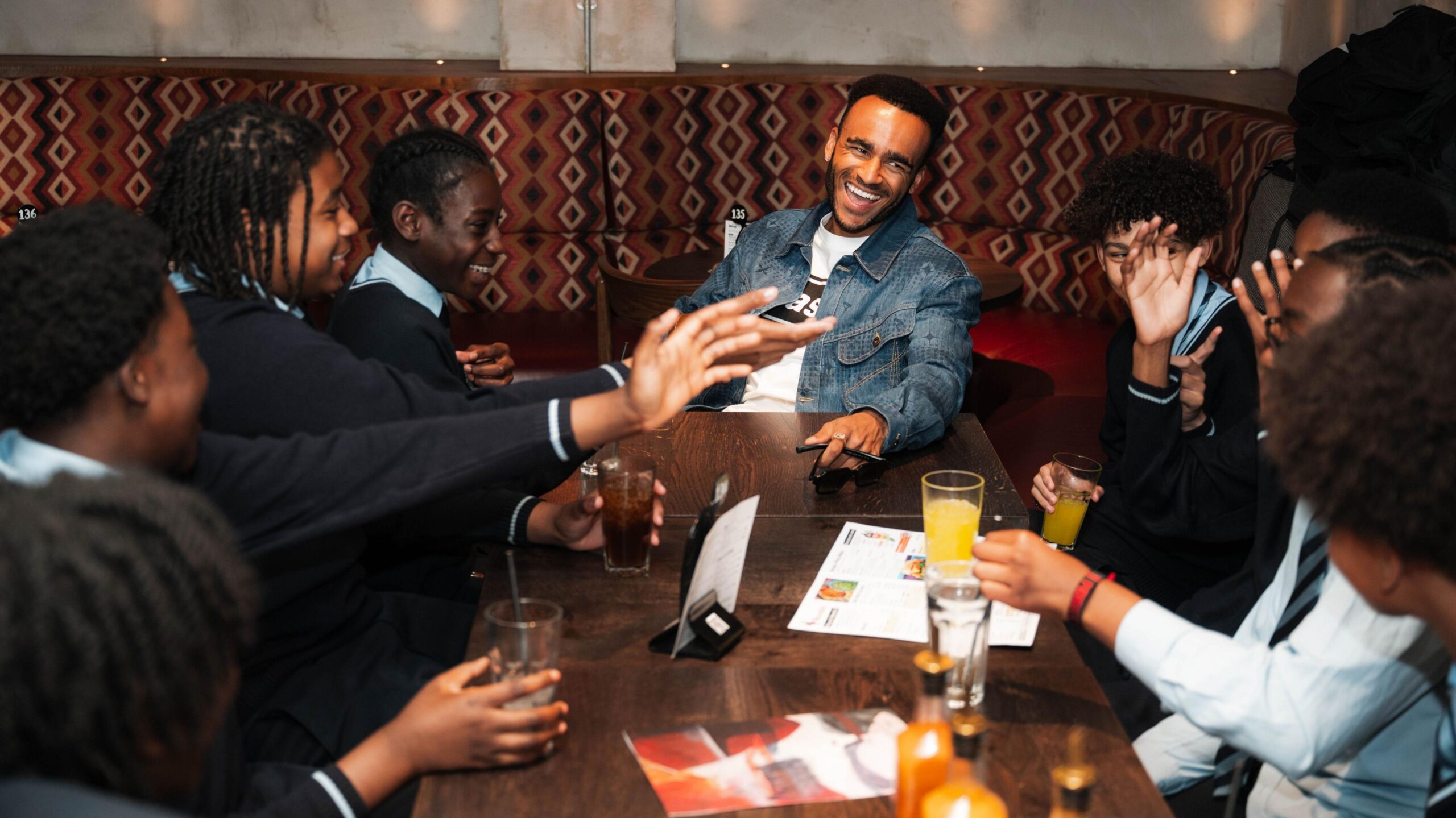 Munya Chawawa sitting and laughing at the head of a table with teenage boys sitting down either side and smiling and talking