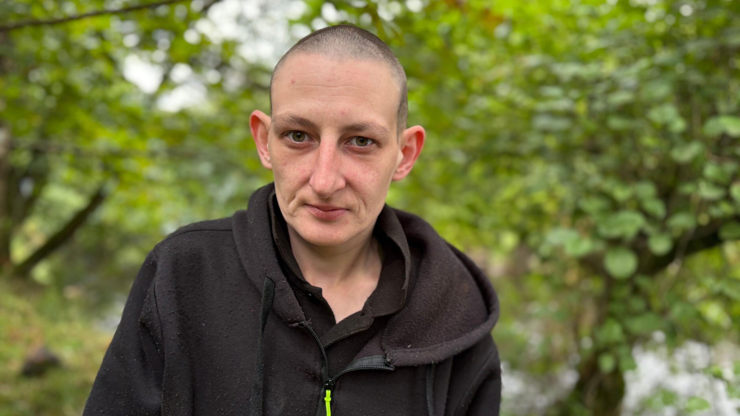 Portrait of a person with closely cropped hair wearing a black hooded top, standing outdoors in a wooded area. Trees and green foliage fill the blurred background, with a stream or body of water visible behind them. The photograph is taken at close range with the subject facing the camera.