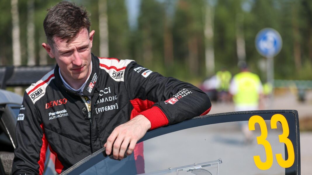 Elfyn Evans in black and red overalls leans on the open door of his number 33 rally car