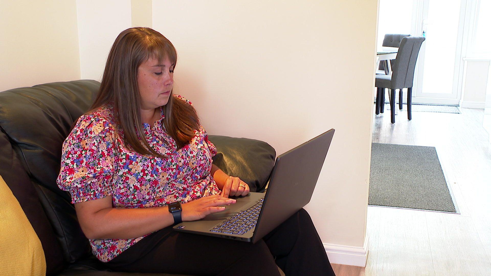 Becky Jones sits on a dark sofa with a laptop open on her lap. She is wearing a brightly flowered blouse and has long dark hair.