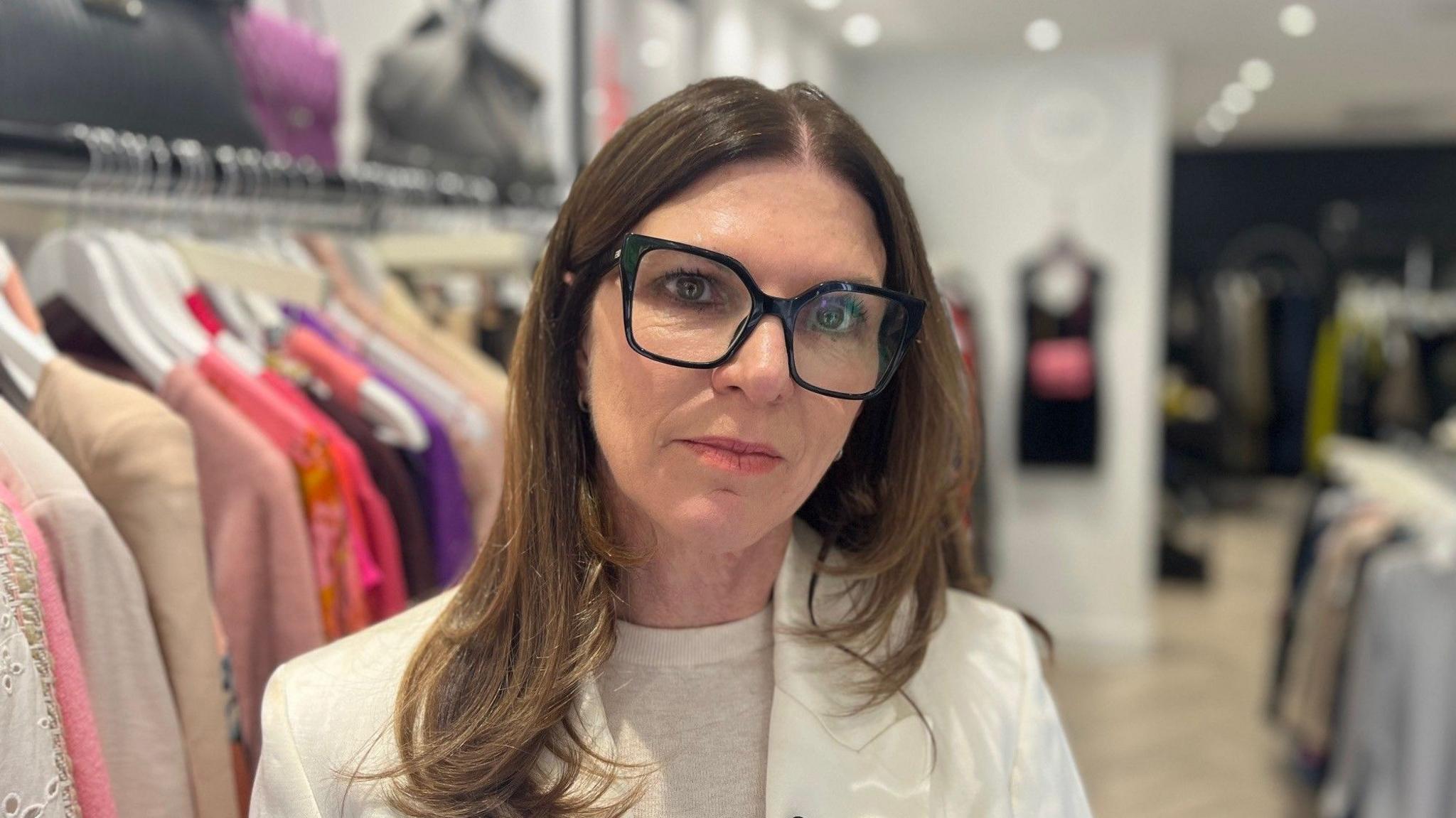 Paige is standing in front of a clothing rack on her shopfloor at her store in Surrey. She is wearing a cream jacket and black glasses.