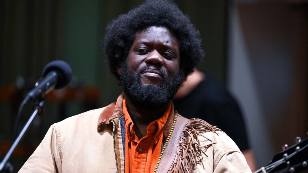 Singer Michael Kiwanuka reveals he is recovering from a stroke Michael Kiwanuka in a light brown jacket and orange shirt, holding a guitar and standing in front of a microphone