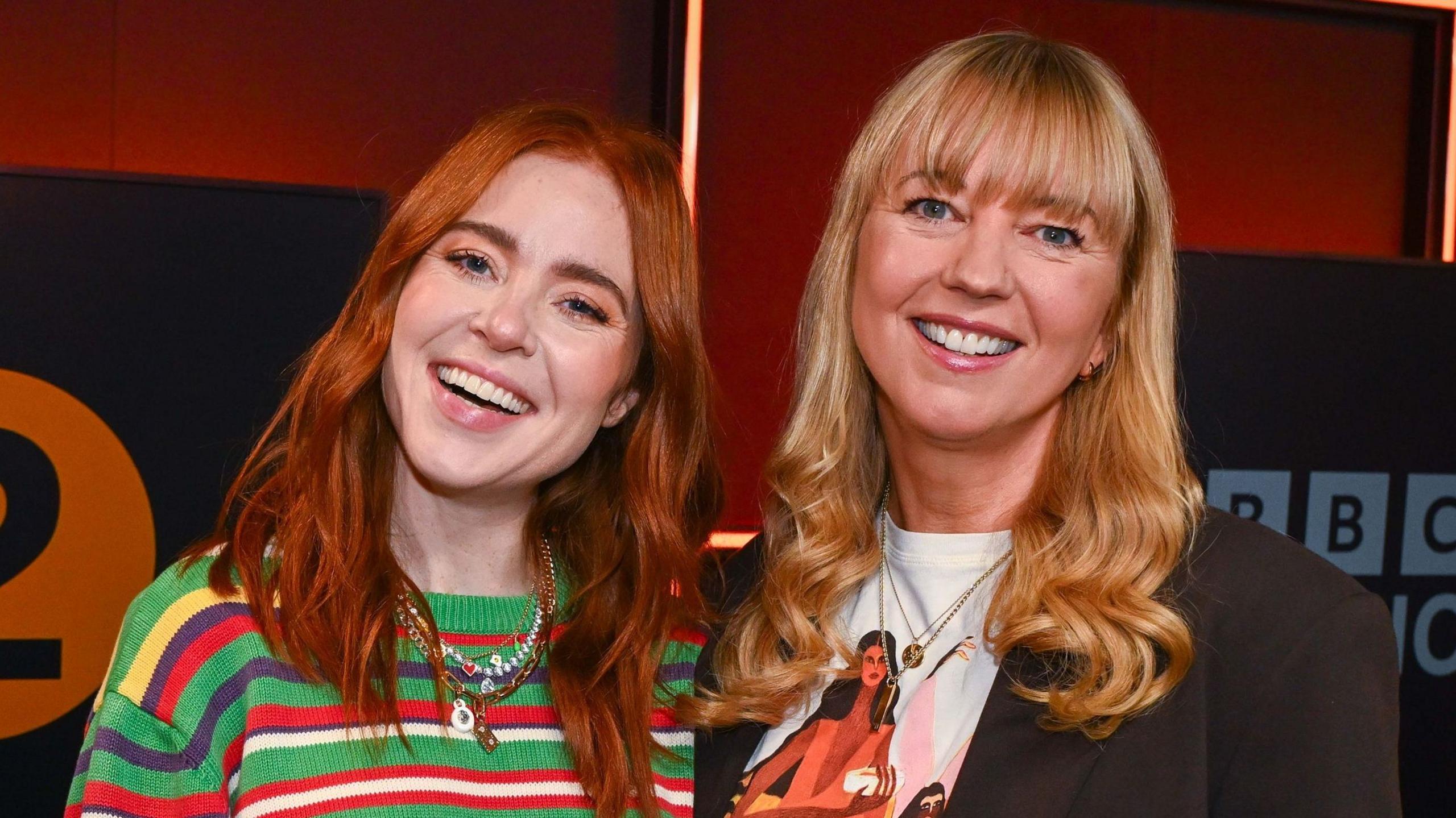 Angela Scanlon and Sara Cox standing and smiling together in front of the BBC Radio 2 logo