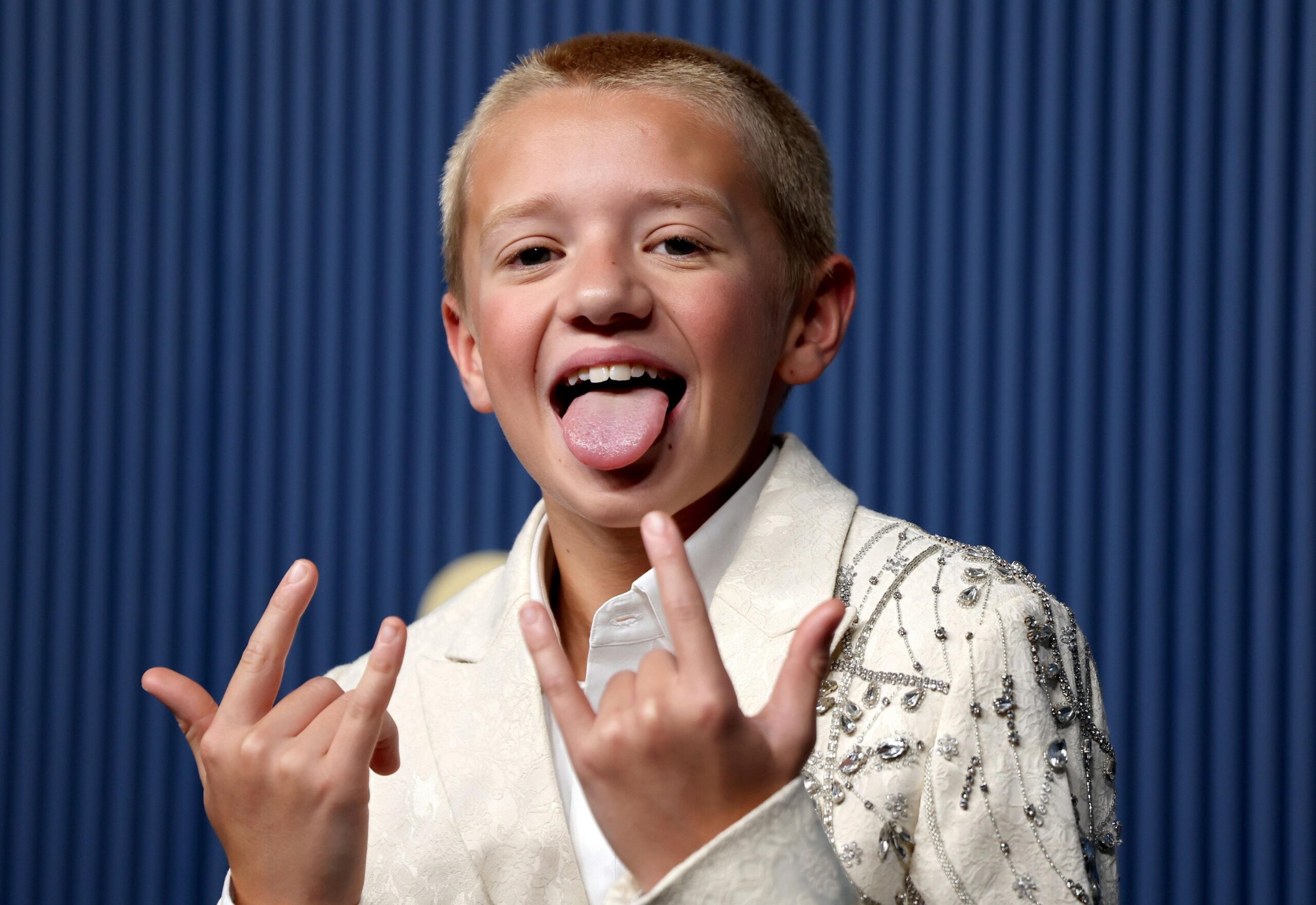 Dexter Sol Ansell in a white jewelled jacket, with his mouth open and tongue out, holding both hands up in the rock sign.