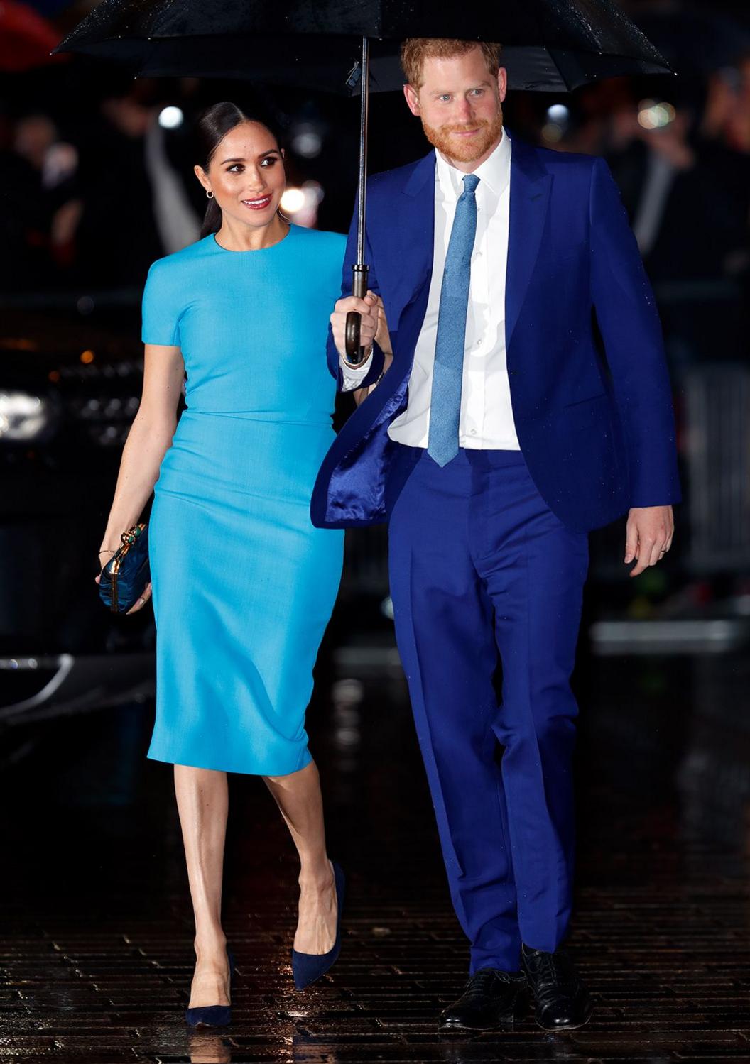 Meghan walks in her dress under an umbrella held by Pince Harry wearing a blue suit, walking outdoors at night.