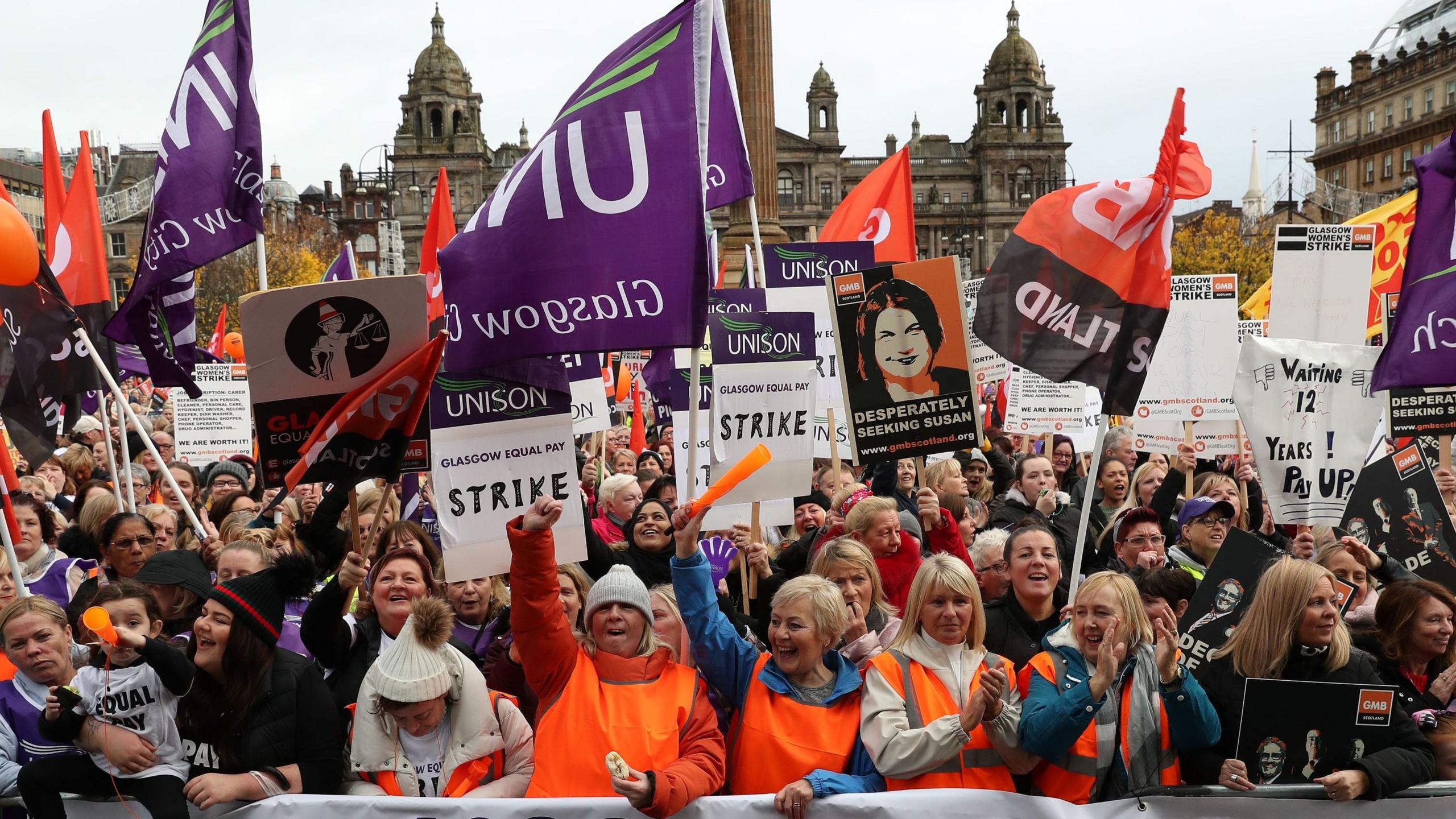 A large group of women stand behind a banner that reads 'Glasgow Equal Pay'. They are wearing orange high visibility vests and standing with their arms raised, shouting and waving Unison flags. In the background is the Glasgow City Council building in George Square.