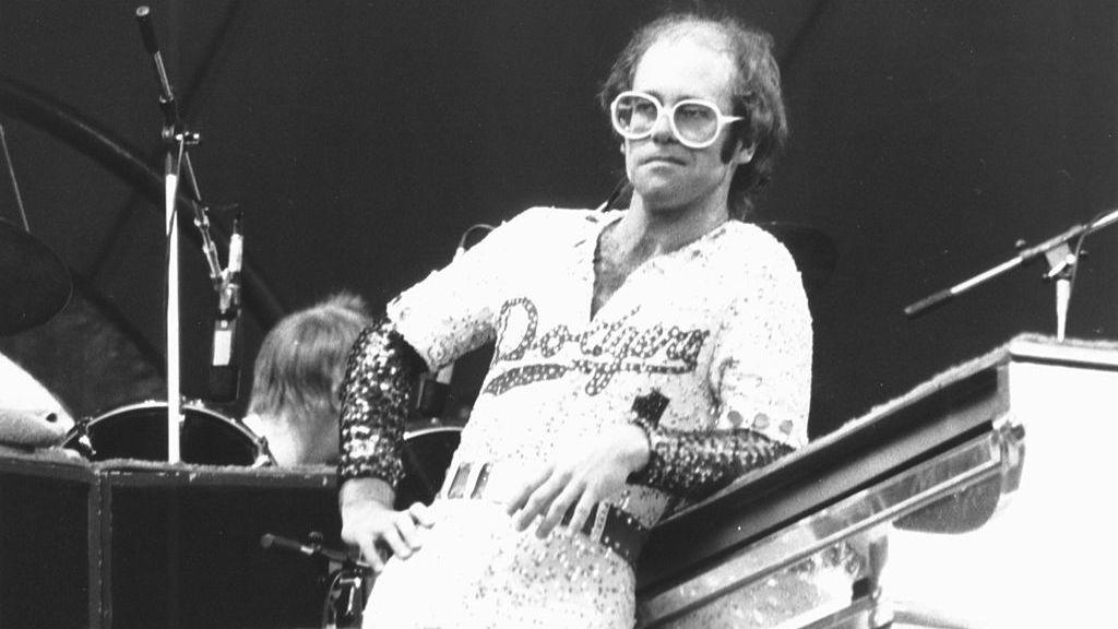 Black and white image shows Elton John leaning against a piano in a bejewelled baseball outfit with 'Dodgers' written across the front.