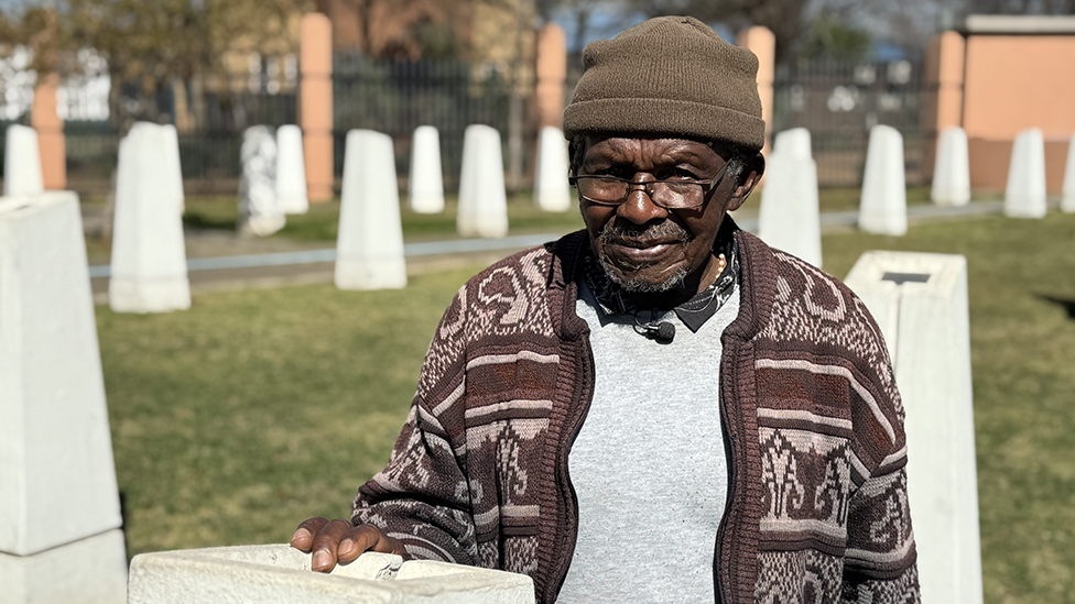 Abraham Mofokeng in a brown patterned cardigan and beanie hat stands by a white monolith. Out of focus, in the background, are other white monoliths - they are part of a memorial.