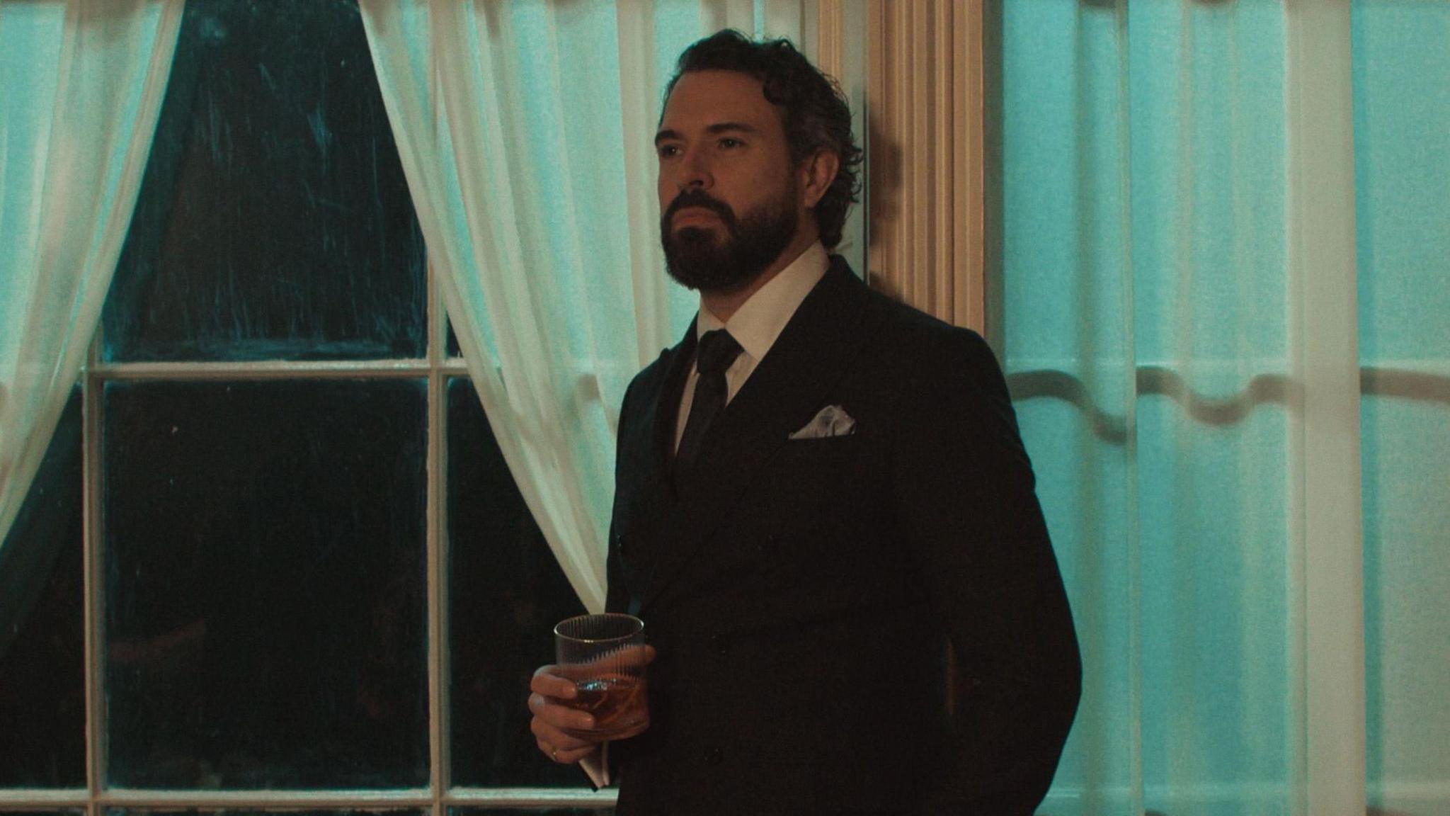 Tom wears a black suit with a white shirt and red tie, He has a grey pocket square. He holds a glass tumbler