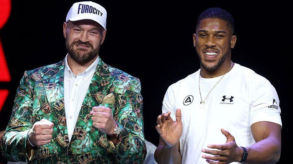Tyson Fury holds up his fists and Anthony Joshua holds up his hands and smiles