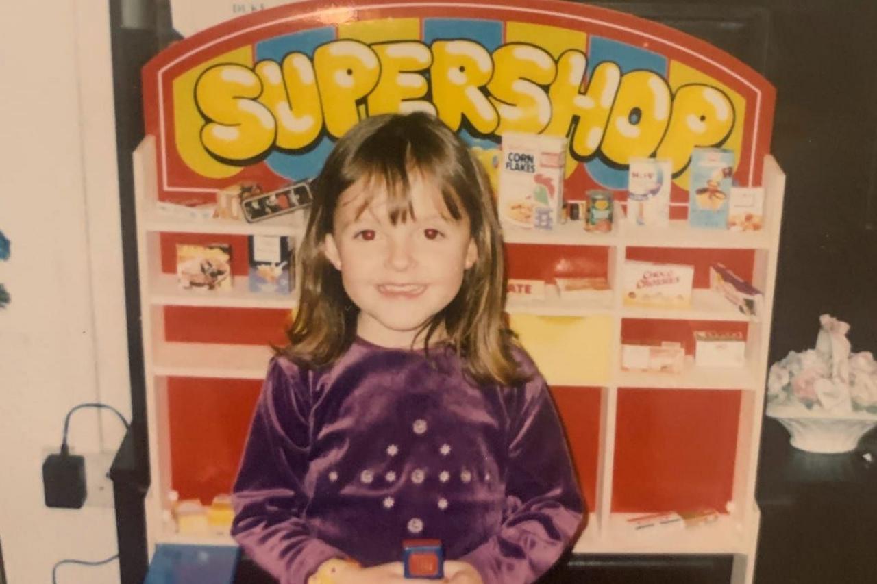 A young child with shoulder-length brown hair and a fringe stands behind a colourful toy checkout and shop display. The child is wearing a long-sleeved purple velvet top decorated with a pattern of small reflective dots across the front and is holding part of the toy checkout equipment with both hands. Behind the child is a red-and-white toy supermarket shelf topped with a large sign reading "SUPERSHOP" in yellow lettering, with miniature grocery boxes and cartons arranged on the shelves, including cereal and biscuit packaging. In the foreground are brightly coloured toy shop accessories, including a blue conveyor-style section, a toy till and a yellow-and-blue scanner. The indoor setting contains additional household items in the background, while the photograph has the slightly soft, warm appearance typical of an older printed family photograph.