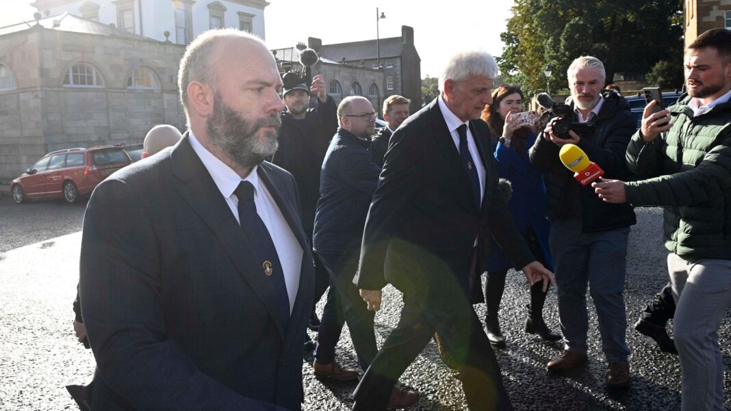 A group of men in suits walking across a carpark, numerous members of the media are holding microphones up to them