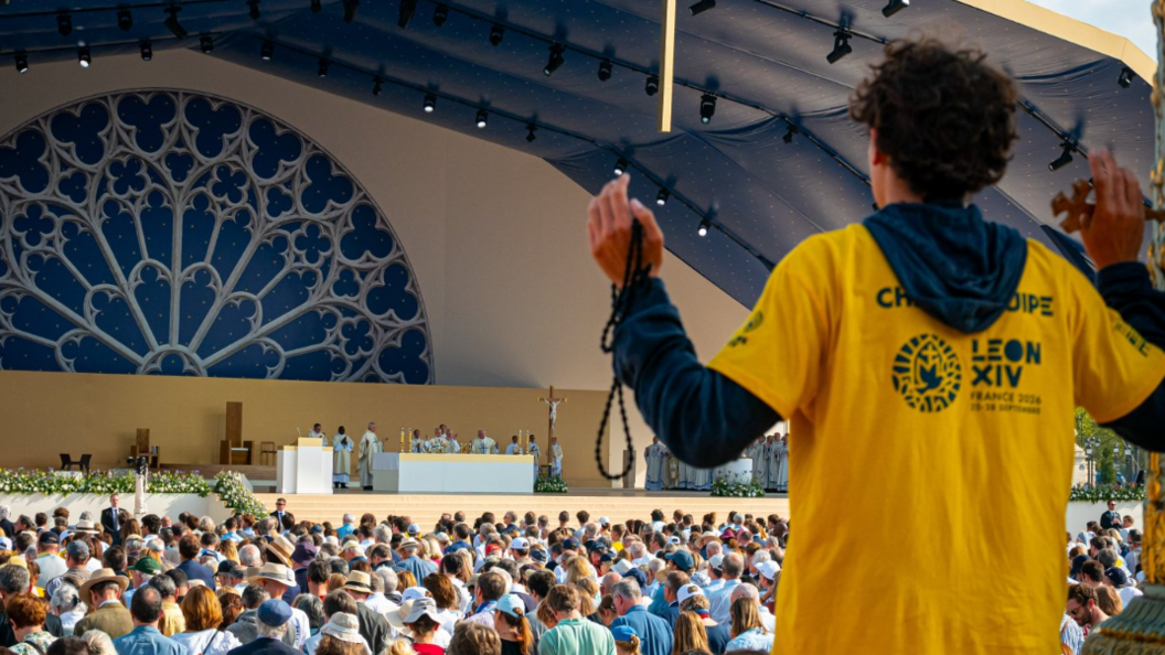 A crowds watches as Pope Leo XIV celebrates Mass on Place de la Concorde, with a young man in a yellow T-shirt with raised arms seen from the back in the foreground