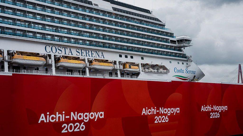 The Costa Serena cruiseliner against cloudy skies, with big boards reading Aichi-Nagoya 2026 in the foreground