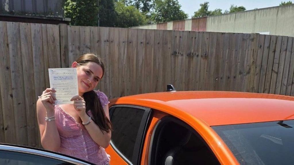 Savannah smiles at the camera holding her pass test certificate for her practical driving test. she has brown hair, is wearing a red top and is stood outside an orange car.