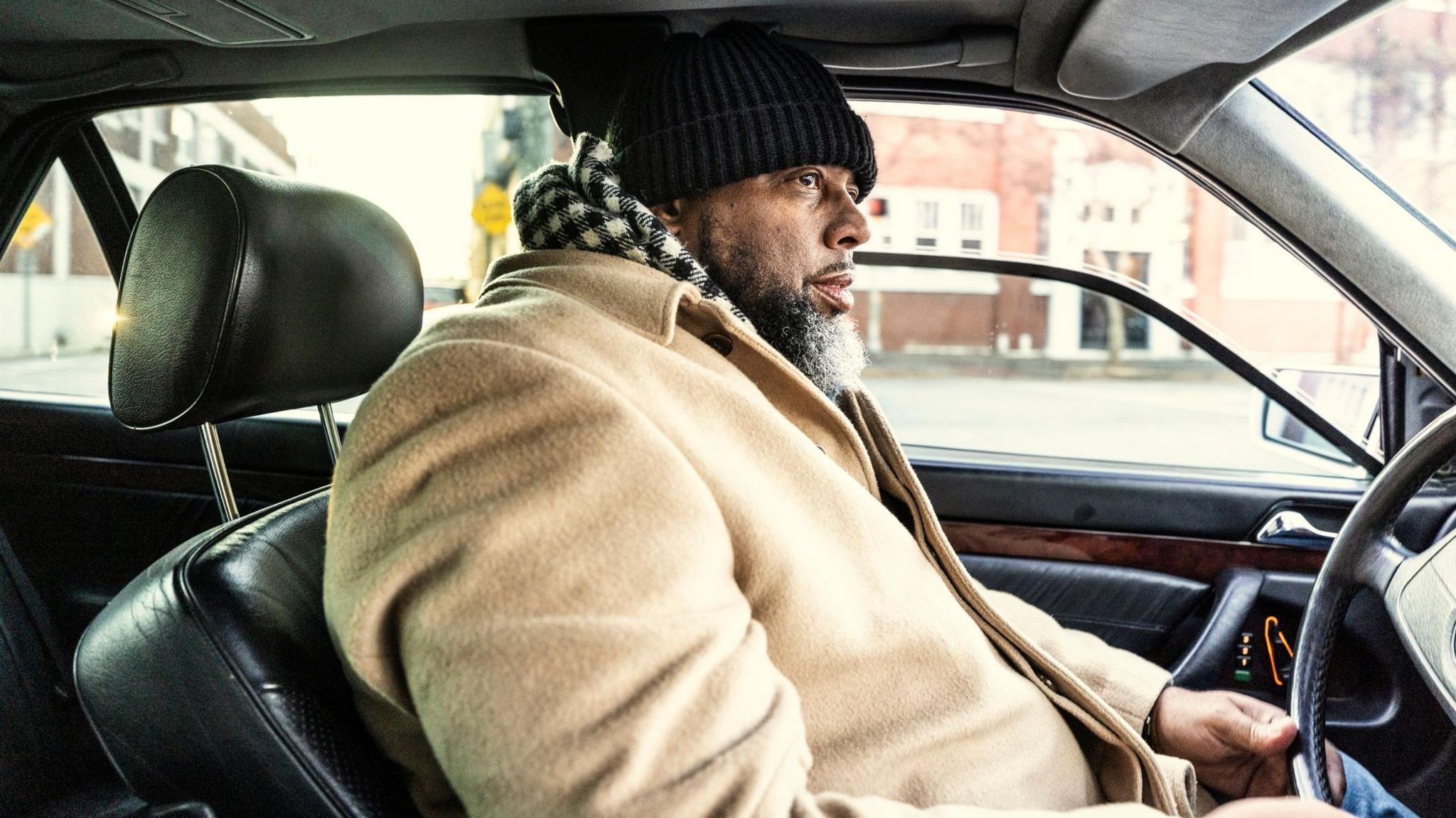 A bearded man in a beige coat and a black and white chequered scarf drives his car through a US urban area. His car has a black leather interior.
