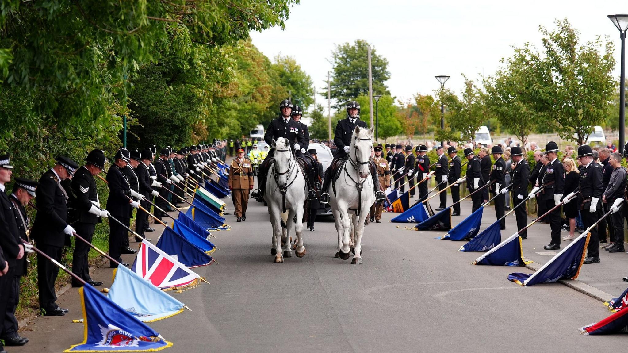 Police officers dressed in all black line the street Tom Clough's coffin is being transported down. The officers have their heads bowed and they are each holding a different flag which is resting on the ground out in front of them. Four mounted officers on horses lead the cortege with multiple black vehicles behind them, along with army soldiers dressed in brown suits and on-duty police officers in yellow hi-vis. There are large trees lining the road with leaves overhanging on the left.