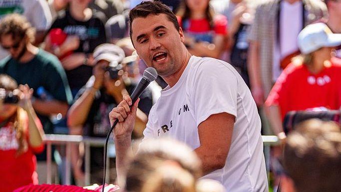 Charlie Kirk hold a microphone and speaks to a crowd of people at a university